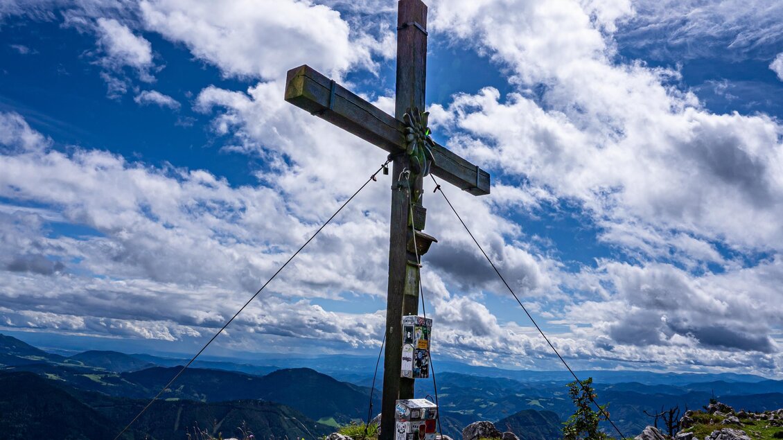 Ein Holzkreuz steht auf einem Berggipfel, umgeben von Felsen und Gras. Der Himmel ist blau mit vereinzelten Wolken und die Sonne strahlt hell. | © Tourismusverband Oststeiermark