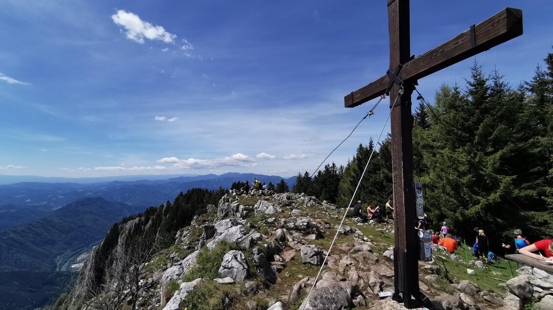 Ein großer Holzkreuz steht auf einem Berggipfel, umgeben von Bäumen. Im Hintergrund erstreckt sich eine weite Aussicht auf die umliegenden Berge und das blaue Himmel. | © Tourismusverband Oststeiermark