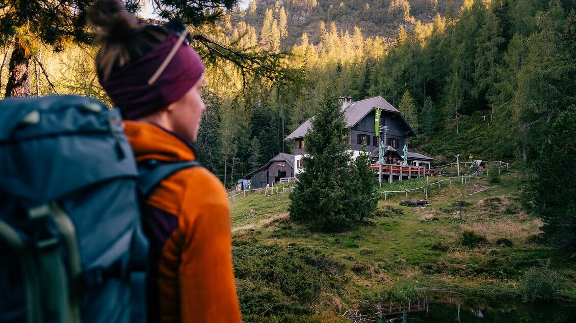 Eine Wanderin mit Rucksack blickt auf eine Berghütte inmitten von Bäumen. Im Hintergrund sind sanfte Hügel und ein klarer Himmel zu sehen.