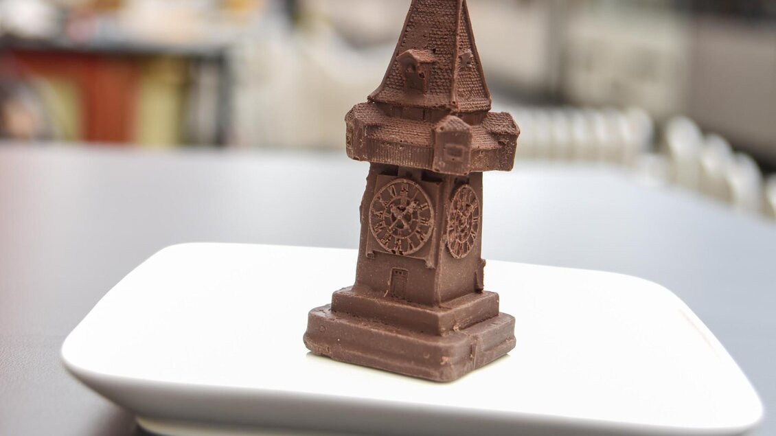 Schokoladenfigur in Form des Grazer Uhrturms auf Teller mit unscharfem Stadtmotiv im Hintergrund. | © Süßer Steirer