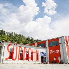 A colorful building with the inscription "Südbahn Museum" on the facade. In the background, forests and a blue sky with white clouds are visible. | © Südbahnmuseum