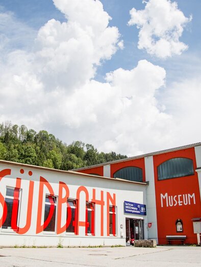 Ein buntes Gebäude mit dem Schriftzug „Südbahn Museum“ an der Fassade. Im Hintergrund sind Wälder und ein blauer Himmel mit weißen Wolken sichtbar. | © Südbahnmuseum