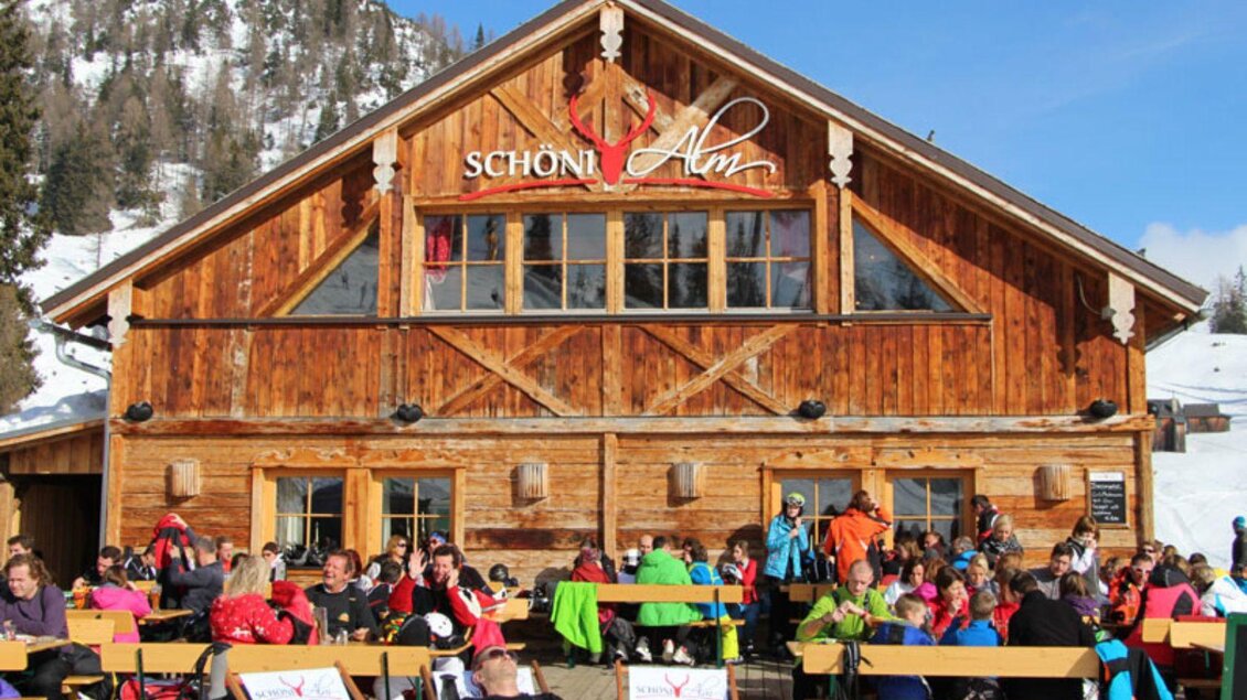 Die Schöni-Alm, eine urige Berghütte mit Holzfassade und vielen Gästen auf der Sonnenterrasse im Freien. Umgeben von schneebedeckten Bergen des Skigebiets Tauplitz genießen die Besucher das schöne Wetter bei einem wunderbaren Skitag. | © Schöni Alm