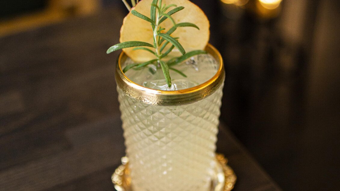 Ein eleganter Cocktail in einem dekorativen Glas auf einem Tisch. Der Drink ist mit einem Rosmarinzweig und einer Zitronenscheibe garniert. | © STIN OG