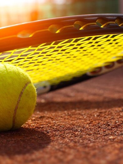 A tennis ball lies on the court next to a tennis racket. The sun shines in the background, illuminating the scene. | © Tennis_AdobeStock_94802234