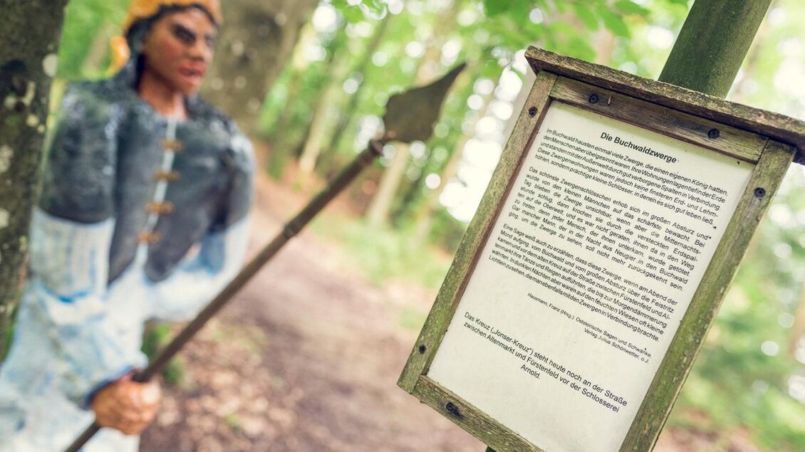 Eine Statue eines Kriegers steht im Wald, daneben befindet sich ein Schild mit Informationen. Die Szene ist umgeben von grünen Bäumen und natürlichem Licht.