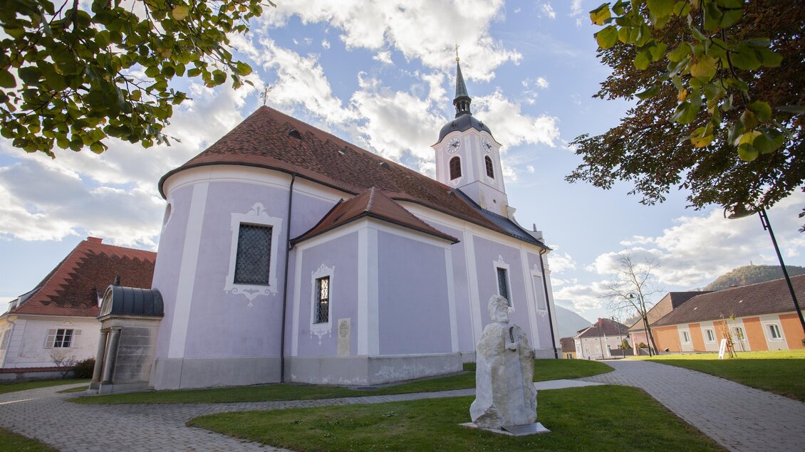 Eine schöne Kirche mit lila Fassade und einem hohen Turm. Umgeben von einer grünen Wiese und unter einem blauen Himmel mit Wolken. | © Tourismusverband Oststeiermark