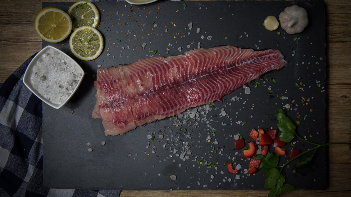 Ein frisches Fischfilet liegt auf einem schwarzen Servierbrett. Dazu sind Zitronenscheiben, Knoblauch, Gewürze und frisches Gemüse angerichtet. | © Salzkammergut Edelwels