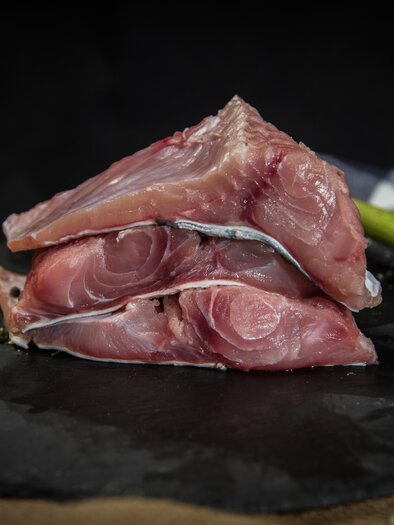 Drei Stücke frischer Fisch liegen auf einem dunklen Teller. Neben dem Fisch sind einige frische Zutaten wie Zitrone und Gemüse zu sehen. | © Salzkammergut Edelwels