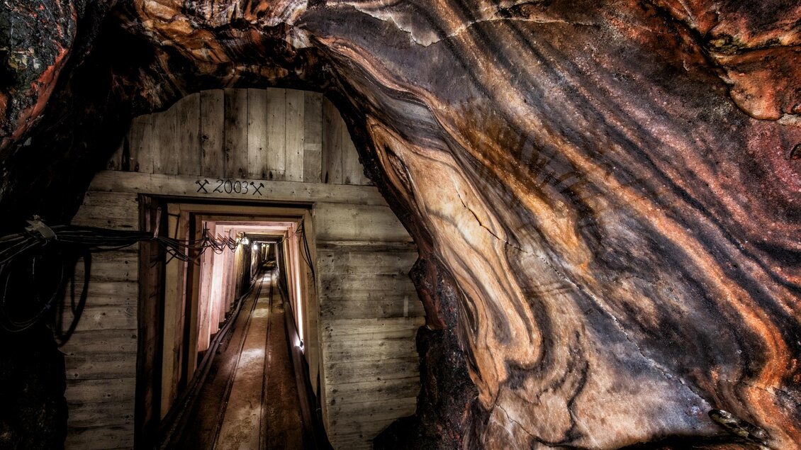 Durch den mit Holz abgestützten Grubenausbau führt der schmale Tunnel ins Innere des Schaubergwerks der Salzwelten in Altaussee. Der Eingang ist umgeben von Wänden aus Steinsalz, welche in den Farben Orange und Schwarz an die Oberfläche treten. | © Salzwelten/Kernmayer
