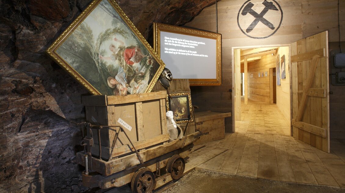 Das Springerwerk im Schaubergwerk der Salzwelten in Altaussee mit einem Förderwagen aus Holz, in dem sich einige Kunstwerke befinden. Im Hintergrund sieht man das Symbol für den Bergbau mit Schlägel und Eisen. | © Salzwelten/Mario Schmid