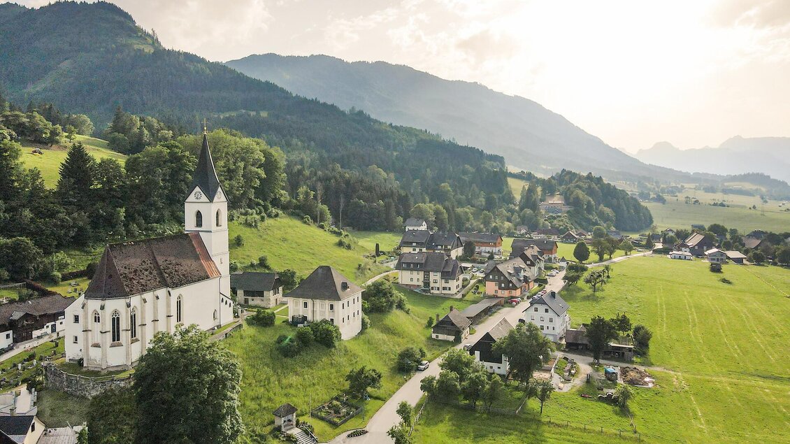 Eine malerische Landschaft mit einer Kirche und kleinen Häusern in einem grünen Tal. Im Hintergrund sind sanfte Hügel und Berge zu sehen.