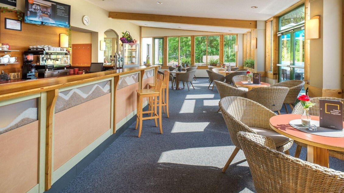 Ein heller Café-Raum mit Holzverkleidung und großen Fenstern. Im Vordergrund stehen gemütliche Rattan-Stühle und Tische, die zum Entspannen einladen. | © JUFA Hotels