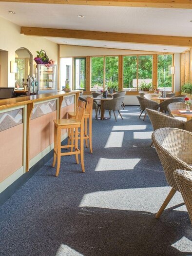 A bright café room with wood paneling and large windows. Cozy rattan chairs and tables in the foreground invite relaxation. | © JUFA Hotels