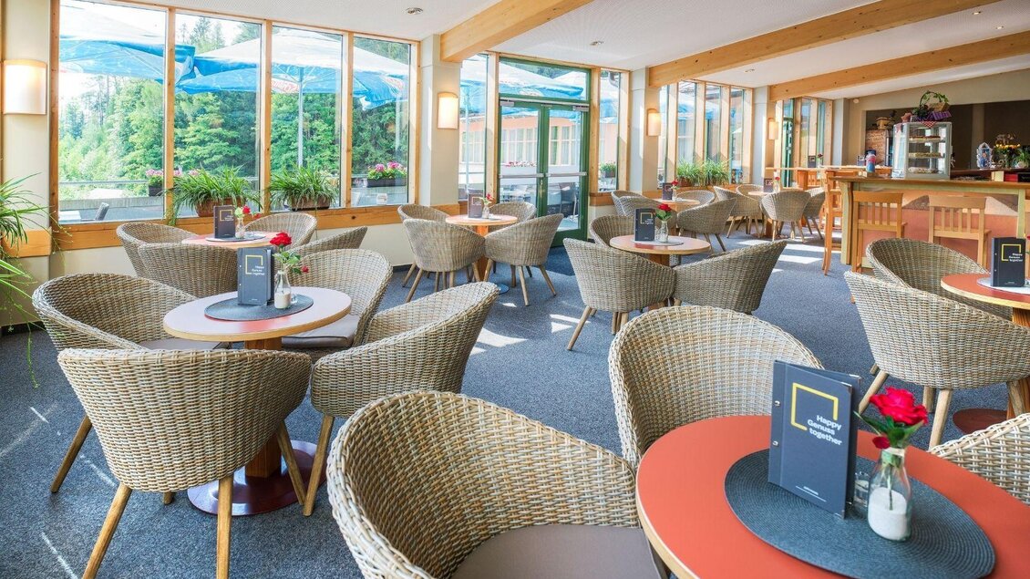 Ein helles Café mit gemütlichen Korbstühlen und Tischen. Große Fenster bieten einen Blick auf die Natur und sorgen für eine einladende Atmosphäre. | © JUFA Hotels
