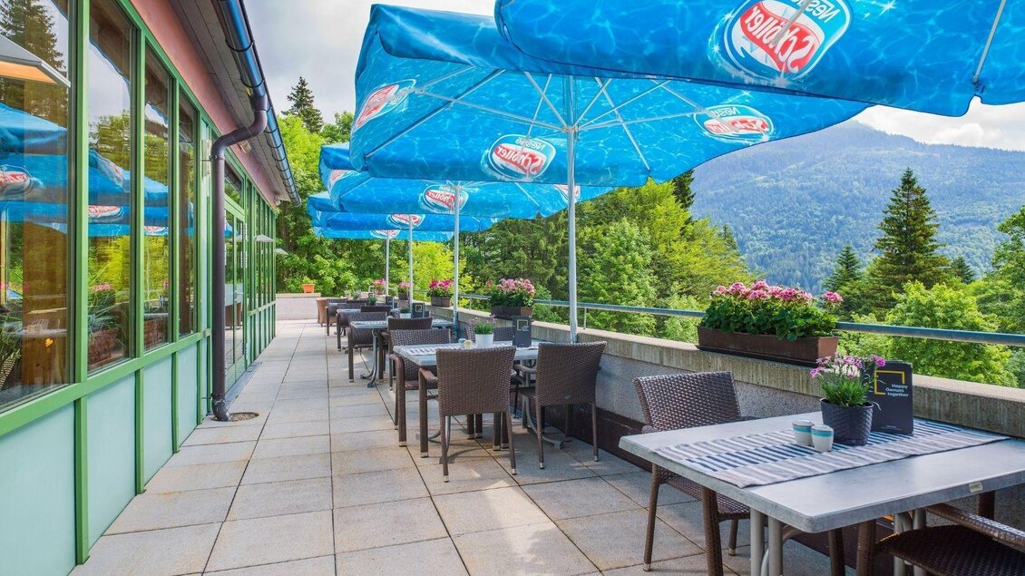 Eine Terrasse mit Tischen und Stühlen, umgeben von grüner Landschaft und Bergen. Große Sonnenschirme bieten Schatten für die Gäste. | © JUFA Hotels