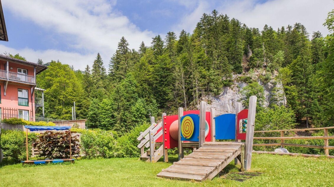 Ein Spielplatz mit einer Rutsche und bunten Spielgeräten, umgeben von grünen Wiesen und Bäumen. Im Hintergrund sind Felsen und eine große Baumlandschaft sichtbar. | © JUFA Hotels