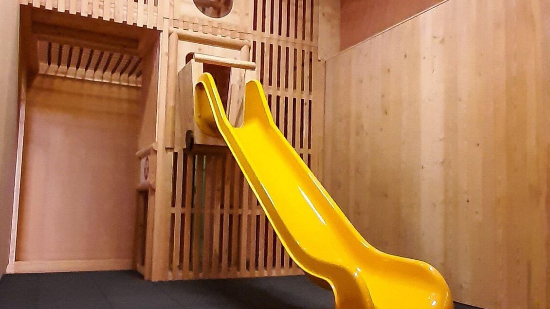 Eine Rutsche aus gelbem Kunststoff in einem Holzspielraum. Die Wände sind aus hellem Holz und der Raum ist gut beleuchtet. | © JUFA Hotels
