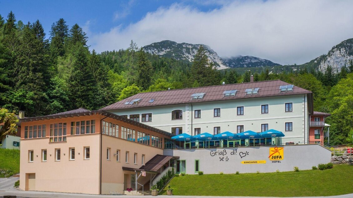 Ein modernes Hotel in bergiger Umgebung mit einem klaren Himmel. Die Fassade bietet schattige Terrassen mit blauen Sonnenschirmen. | © JUFA Hotels