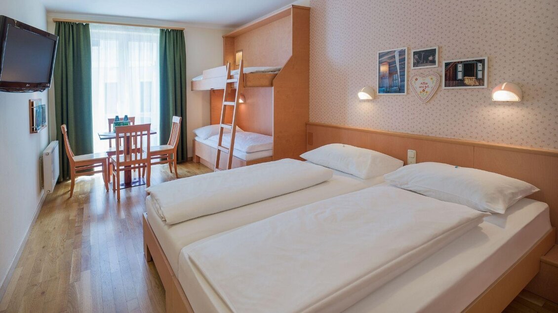 Ein gemütliches Hotelzimmer mit einem Doppelbett und einem Etagenbett. Es gibt ein kleines Esszimmer und große Fenster mit grünen Vorhängen. | © JUFA Hotels
