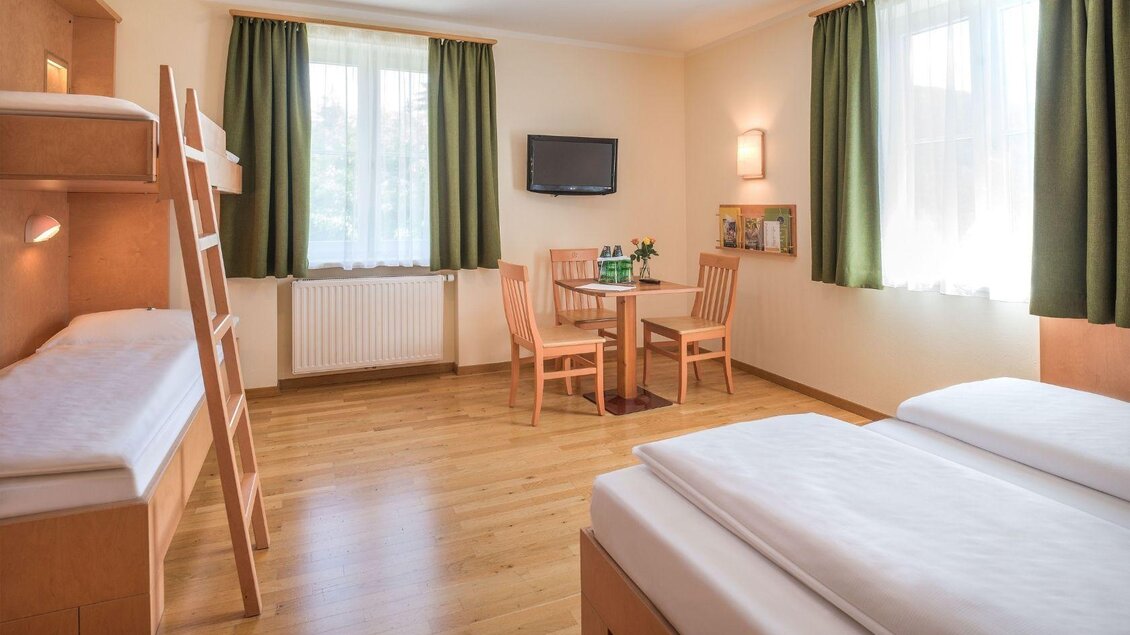 Ein gemütliches Zimmer mit zwei Betten und einem Etagenbett. Ein Tisch mit Stühlen und große Fenster sorgen für viel Licht. | © JUFA Hotels
