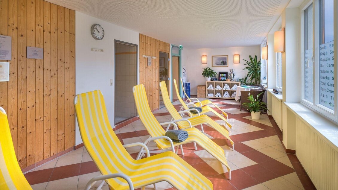 Ein heller Wellnessbereich mit gelben Liegen und großen Fenstern. Pflanzen und eine Uhr schaffen eine entspannte Atmosphäre. | © JUFA Hotels