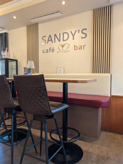 Ein gemütlicher Café-Bereich mit einem Tisch und Stühlen. An der Wand befindet sich das Schild "SANDY'S café bar". | © Sandy`s Café Bar