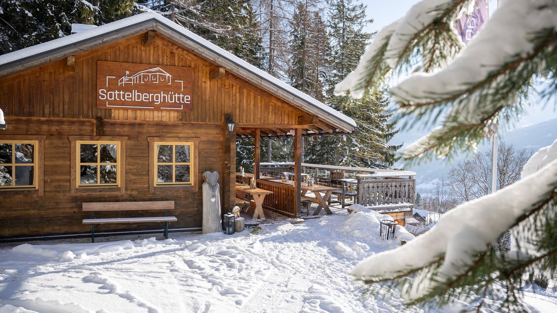 Eine gemütliche Berghütte im Schnee, umgeben von Tannenbäumen. Der klare Himmel und die Sonne verleihen der Szene eine einladende Atmosphäre.