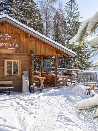 A cozy mountain cabin in the snow, surrounded by fir trees. The clear sky and the sun give the scene an inviting atmosphere.