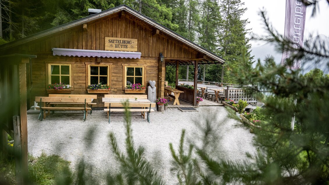 Eine gemütliche Holzsch hut mit Blumenfenstern, umgeben von grünen Bäumen. Draußen stehen Holztische und -bänke für Besucher. | © Sattelberghütte