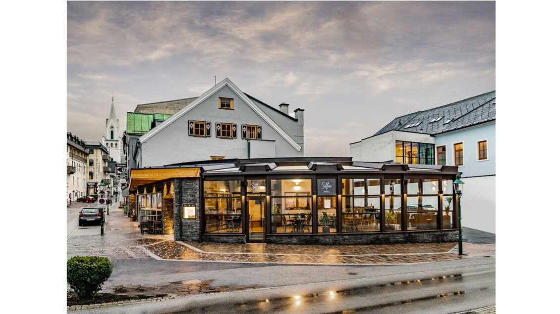 Ein modernes Restaurant mit großen Fenstern in einer malerischen Stadt. Die Umgebung ist ruhig mit einigen Gebäuden und einem blauen Himmel im Hintergrund. | © Christine Höflehner