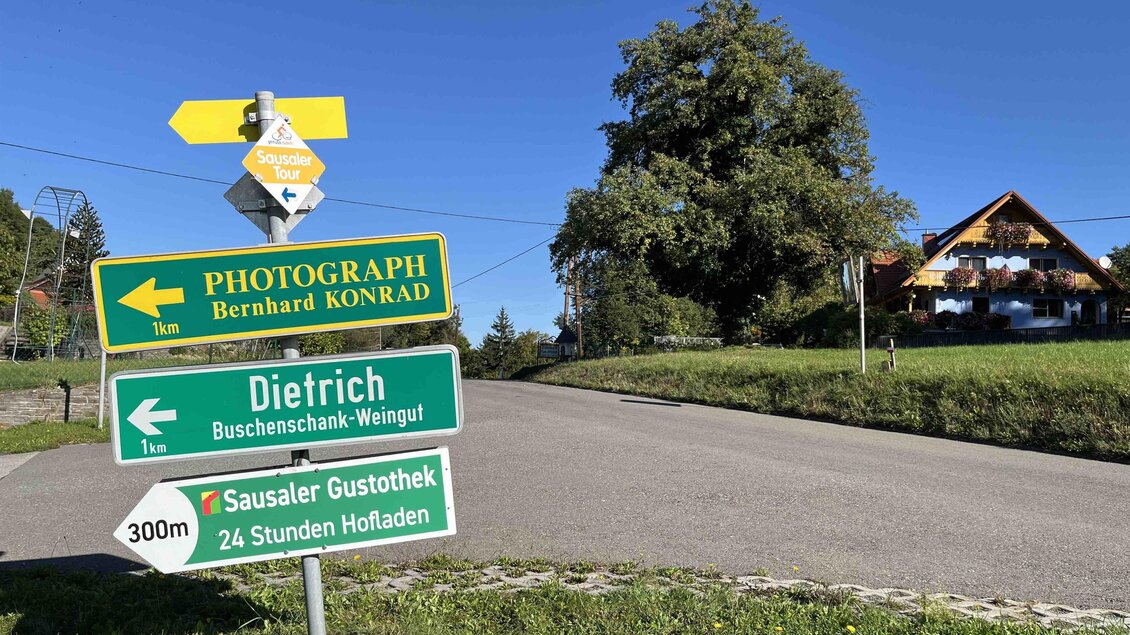 Eine Straße mit mehreren Wegweisern und einem Gebäude im Hintergrund. Die Richtungsschilder zeigen zu verschiedenen Zielen wie einem Weingut und einer Gaststätte. | © Sausaler Gustothek | Dr. Ulrike Schauer