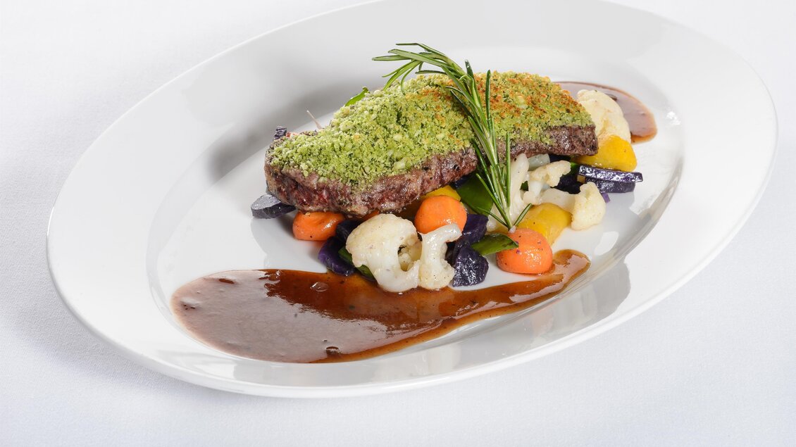 Ein zartes Stück Fleisch, überzogen mit einer Kräuterkruste, liegt auf buntem Gemüse. Daneben befindet sich eine dunkle Sauce auf dem Teller. | © Tauroa GmbH