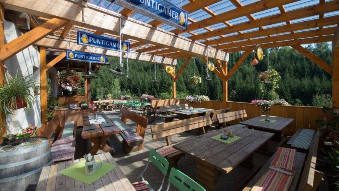 Ein gemütlicher Außenbereich eines Restaurants mit Holztischen und Bänken. Die Umgebung wird von Pflanzen und einem schönen Blick auf die Natur umgeben.
