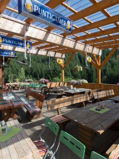 A cozy outdoor area of a restaurant with wooden tables and benches. The surroundings are filled with plants and a beautiful view of nature.