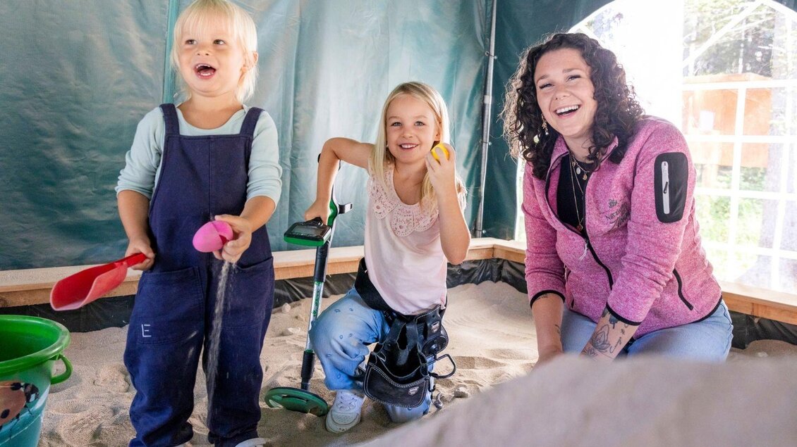 Zwei Kinder spielen im Sandkasten und lächeln glücklich. Eine Frau sitzt neben ihnen und wirkt ebenfalls fröhlich. | © www.zloam.at
