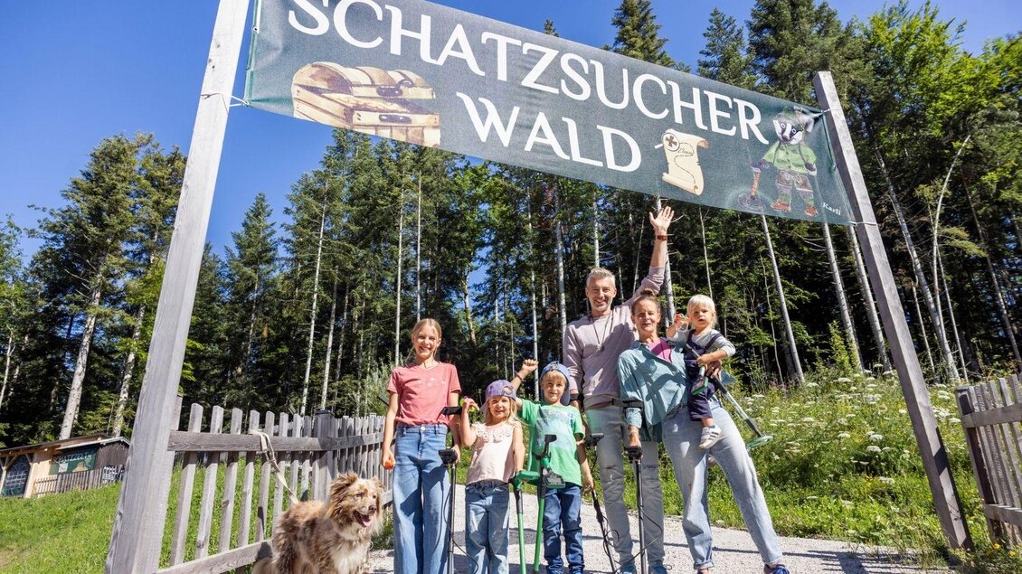 Eine Familie steht vor dem Eingang zu einem Wald mit einem Schild, das "Schatzsucher Wald" zeigt. Sie sind umgeben von Bäumen und genießen einen sonnigen Tag.
