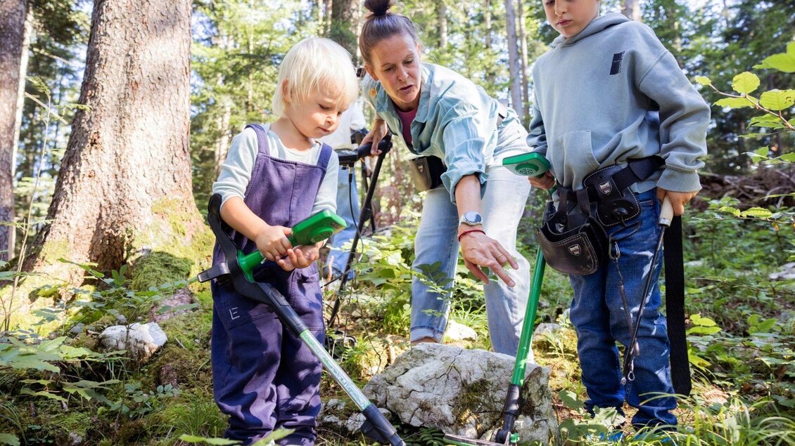 Eine Frau zeigt zwei Kinder, wie man mit einem Werkzeug in der Natur arbeitet. Sie stehen in einem bewaldeten Gebiet mit vielen Pflanzen und Bäumen.