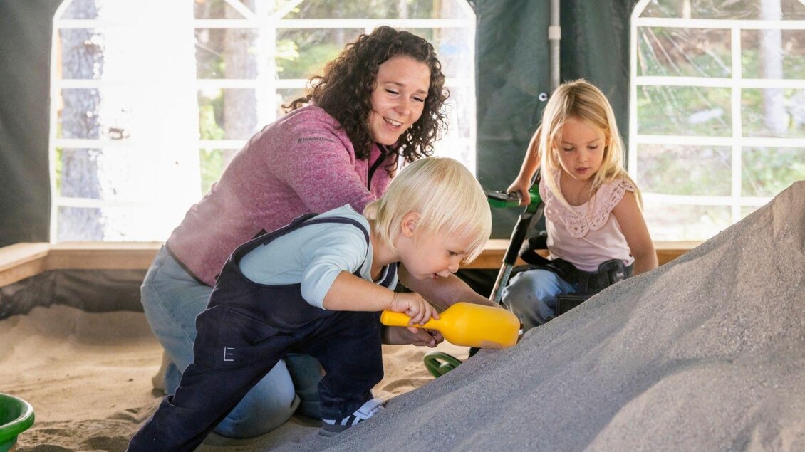 Eine Mutter spielt mit ihren Kindern im Sandkasten. Die Kinder zeigen Freude beim Spielen mit Schaufeln und Sand. | © www.zloam.at