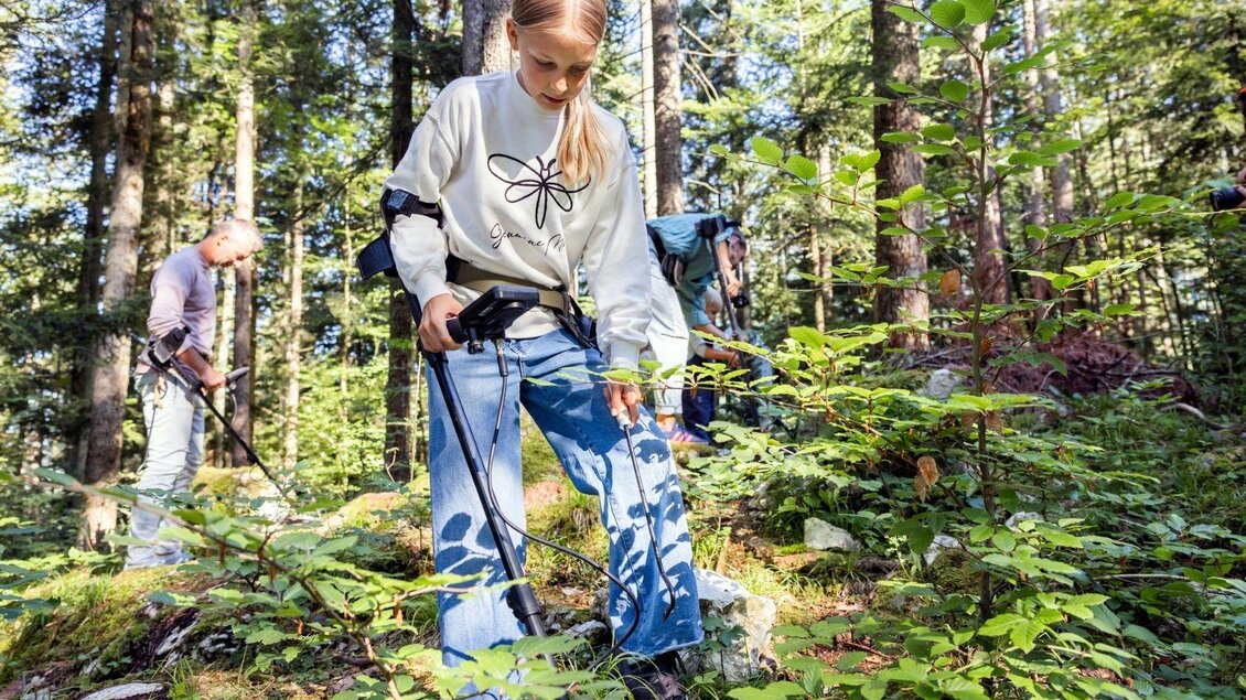 Eine Gruppe von Personen sucht im Wald mit Metalldetektoren. Sie stehen zwischen Bäumen und Pflanzen und sind auf der Suche nach verborgenen Objekten.