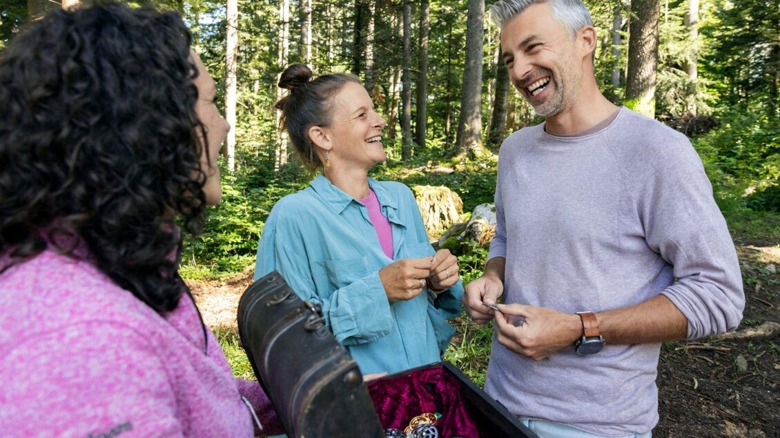 Drei Freunde lachen und unterhalten sich im Wald, während sie gemeinsam einen Schatz aus einer Kiste betrachten. Die angenehme Atmosphäre und der grüne Hintergrund schaffen eine fröhliche Stimmung.