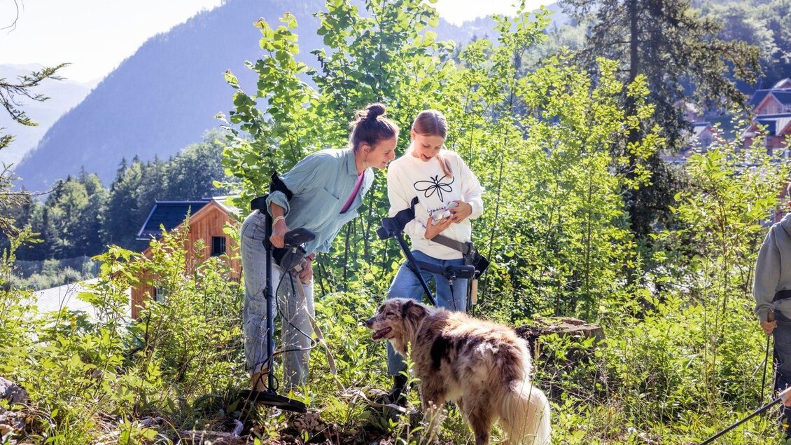 Zwei Frauen arbeiten im Freien und untersuchen den Boden. Ein Hund steht neben ihnen inmitten von Bäumen und Grün. | © www.zloam.at