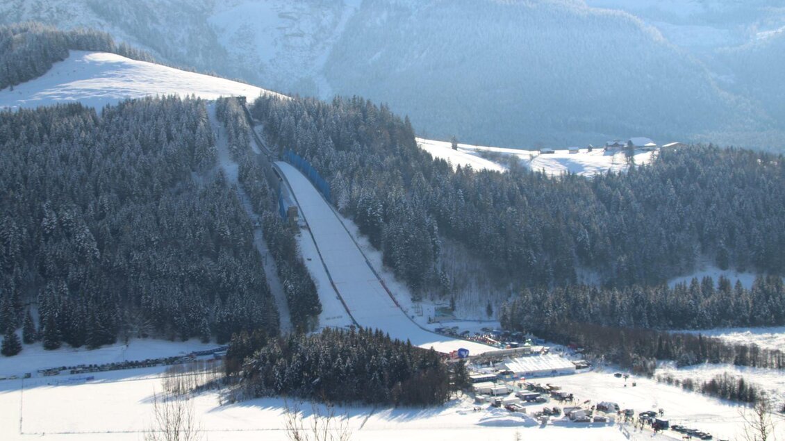 <p>Eine schneebedeckte Landschaft mit der Skiflugschanze, Kulm vor einem bewaldeten Hügel. Im Hintergrund ist der Grimming und ein klarer Himmel zu sehen.</p> | © TVB Ausseerland Salzkammergut/Kolb