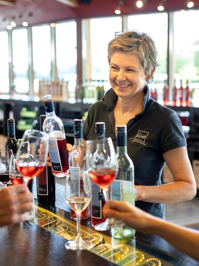 Luise Jöbstl lächelt, während sie Gästen Weinproben zum Verkosten einschänkt. Im Hintergrund sind große Fenster und eine ansprechende Weinpräsentation zu sehen.