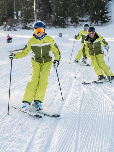 Eine belebte Skipiste mit vielen Skifahrern und farbenfrohen Skianzügen. Im Hintergrund sind Wälder und ein bewölkter Himmel zu sehen. | © Klaus Ranger