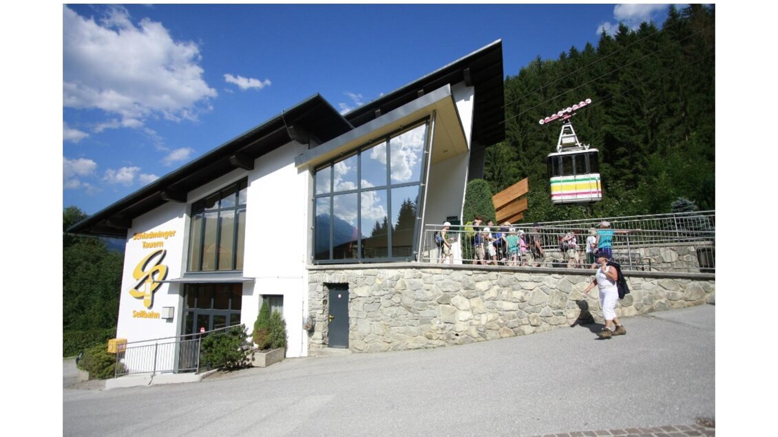 Ein modernes Bergbahngebäude mit großen Fenstern und einer wartenden Menschenmenge. Im Hintergrund ist eine Gondelbahn zu sehen, umgeben von grünen Wäldern und blauem Himmel. | © Verein Schaferlebnis am Hauser Kaibling