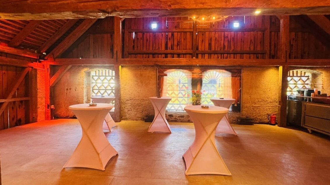 Eine rustikale Veranstaltungsstätte mit Holzdecke und warmem Licht. Mehrere Stehtische stehen im Raum, umgeben von alten Ziegelwänden und großen Fenstern. | © Hannes Kaufmann