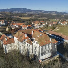 A large, unfinished building with red roofs in a rural setting. In the background, gentle hills and more houses are visible. | © Schlösserstraße