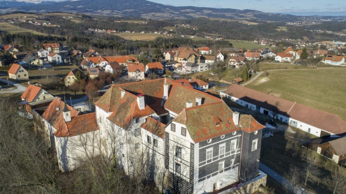 Ein großes, unvollendetes Gebäude mit roten Dächern in einer ländlichen Umgebung. Im Hintergrund sind sanfte Hügel und weitere Wohnhäuser zu sehen. | © Schlösserstraße