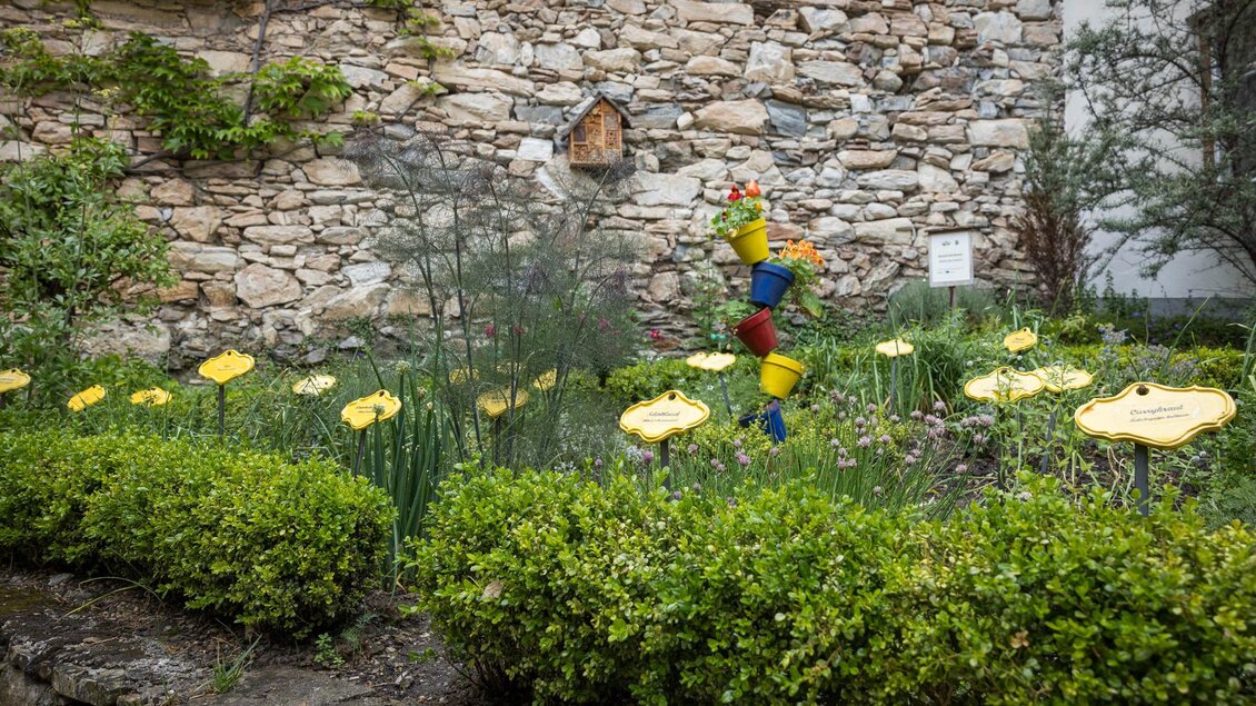 Ein bunter Garten mit bunten Blumentöpfen und gelben Schildern. Im Hintergrund ist eine steinerne Wand und grüne Sträucher zu sehen. | © wildbild